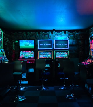 Dimly lit gaming arcade featuring a variety of brightly illuminated slot machines and video gaming screens. Neon signs and colorful displays create a vibrant atmosphere. The arrangement of gaming machines suggests an inviting space for entertainment, with chairs positioned for players.