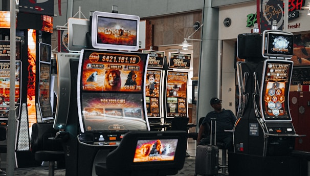 A collection of slot machines is situated inside a building with bright, colorful screens displaying various games and gaming themes. A person is seated nearby, seemingly relaxed with luggage beside them, and the atmosphere is lively with electronic displays.